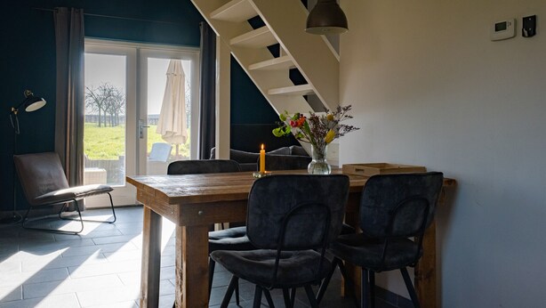Uma área de refeições com mesa de madeira, cadeiras escuras, um vaso com flores, uma luminária pendente e vista através de uma porta de vidro.