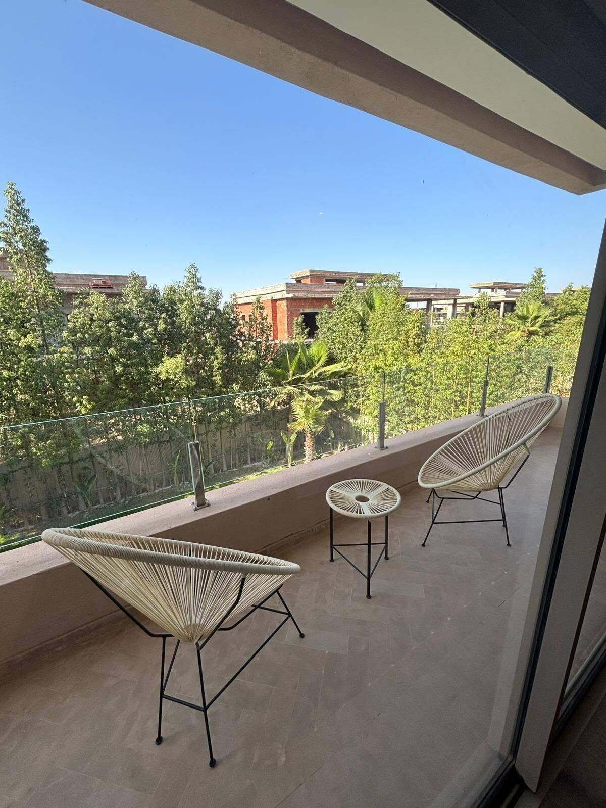 A balcony with wicker chairs and a small table, overlooking a residential area with trees and buildings.