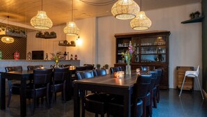 Un comedor con varias mesas y sillas, lámparas de mimbre colgantes, un aparador de madera con puertas de cristal y una pequeña silla blanca.