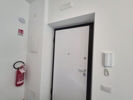 Una puerta blanca con marco negro, un extintor, un portero automático en la pared y un interruptor de luz.