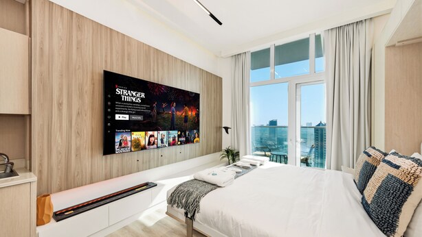 Una moderna camera d'albergo con un'ampia televisione a parete, un letto con biancheria da letto bianca, un piccolo angolo salotto e vista sulla città attraverso ampie finestre.