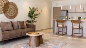 Una sala moderna con un sofá, una mesa redonda de centro y una planta. Cuenta con una zona de cocina con un refrigerador, un lavamanos y taburetes de bar.