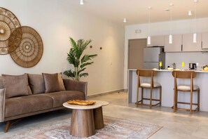 Ein modernes Wohnzimmer mit einer Couch, einem runden Couchtisch und einer Pflanze. Es gibt eine Küchenzeile mit einem Kühlschrank, einer Spüle und Barhocker.