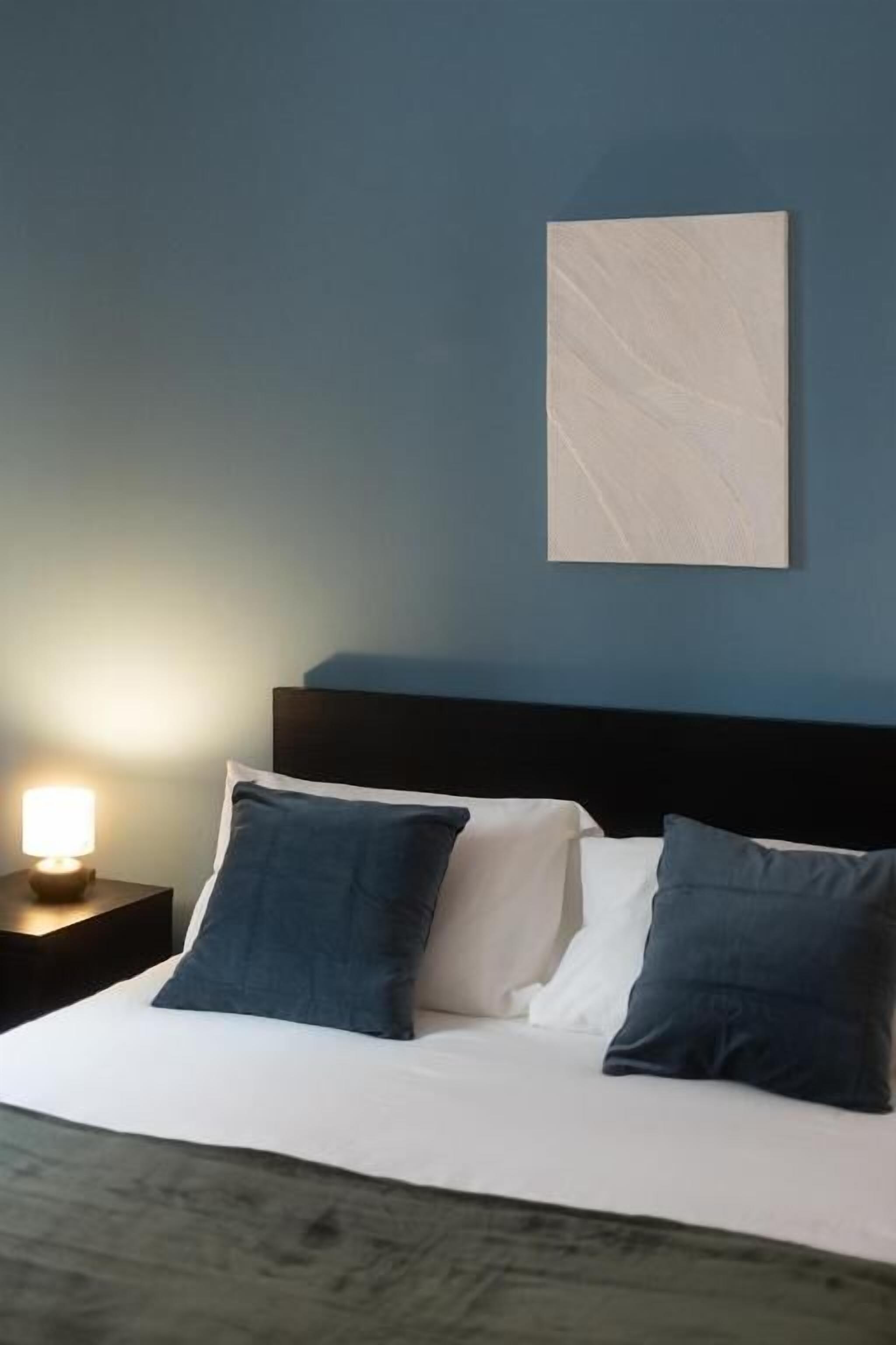 Ein Bett mit weißer und dunkelblauer Bettwäsche, eine Nachttischlampe und ein Wandbild.