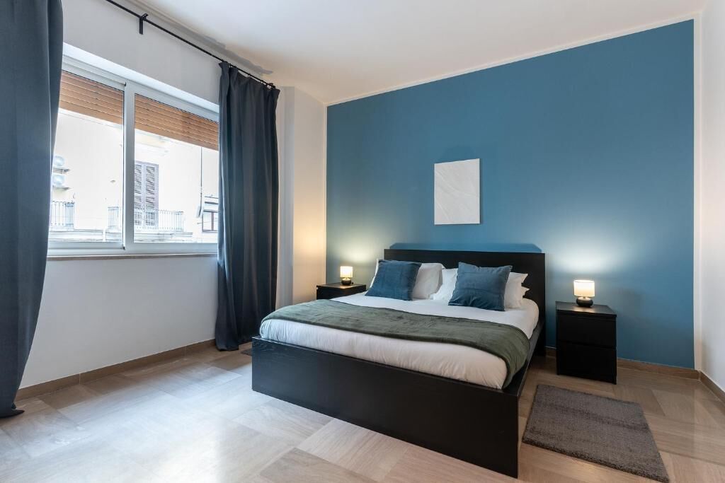 Un dormitorio con cama, dos mesitas de noche, una ventana con cortinas y una pared azul.