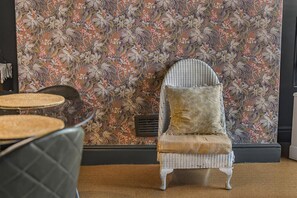 A wicker chair with a cushion, a patterned floral wallpaper, and a vent on the wall. - Guestready - Rose and Monkey (Manchester)