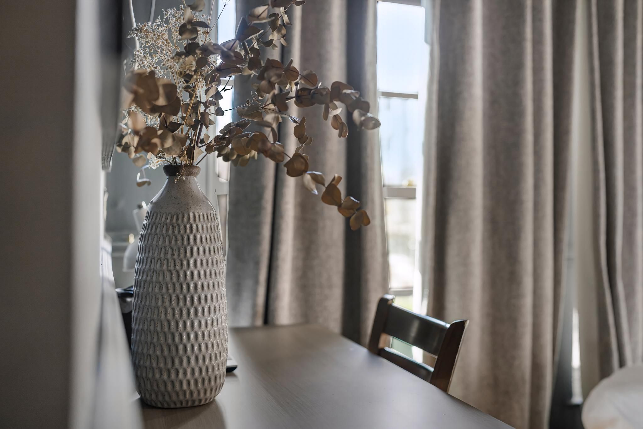 A dining table with a vase of dried flowers, a chair, and curtains.