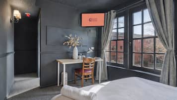 Quarto de hotel com uma cama, uma escrivaninha com cadeira, um vaso com flores, uma televisão na parede e uma janela com cortinas.
