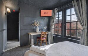 A hotel room with a bed, a desk with a chair, a vase with flowers, a wall-mounted TV, and a window with curtains. - Guestready - Rose and Monkey (Manchester)