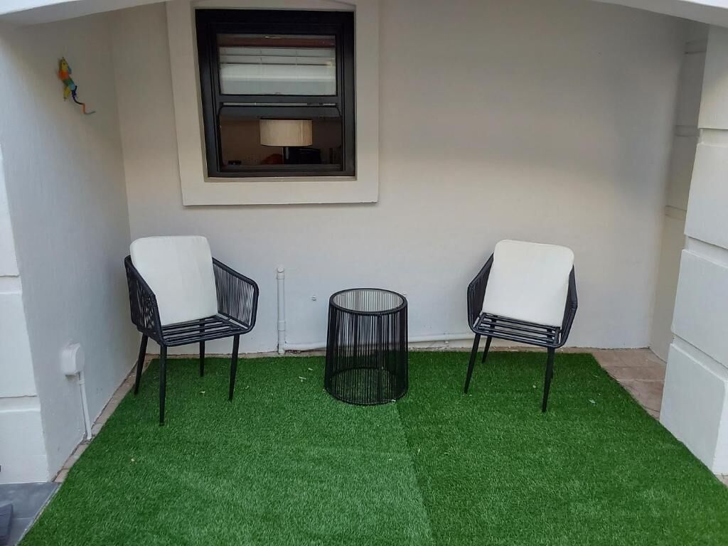 A small outdoor area with two chairs and a round table, a window, and artificial grass.