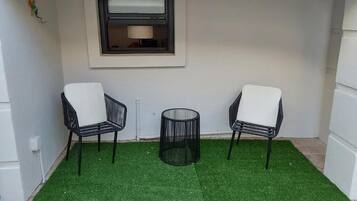 A small outdoor area with two chairs and a round table, a window, and artificial grass.