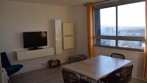 Une chambre moderne dotée d’une télévision à écran plat, d’une table à manger avec des chaises, d’une fenêtre avec des rideaux et offrant une vue sur la ville.