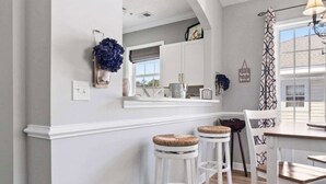 A kitchen with white cabinetry, a white countertop, and a wooden floor. There are three white stools with cushioned seats. - Barefoot Bliss-Updated 2 Bedroom, 2 Bathroom Retreat! Beach, Golf, Pools, More! (North Myrtle Beach)