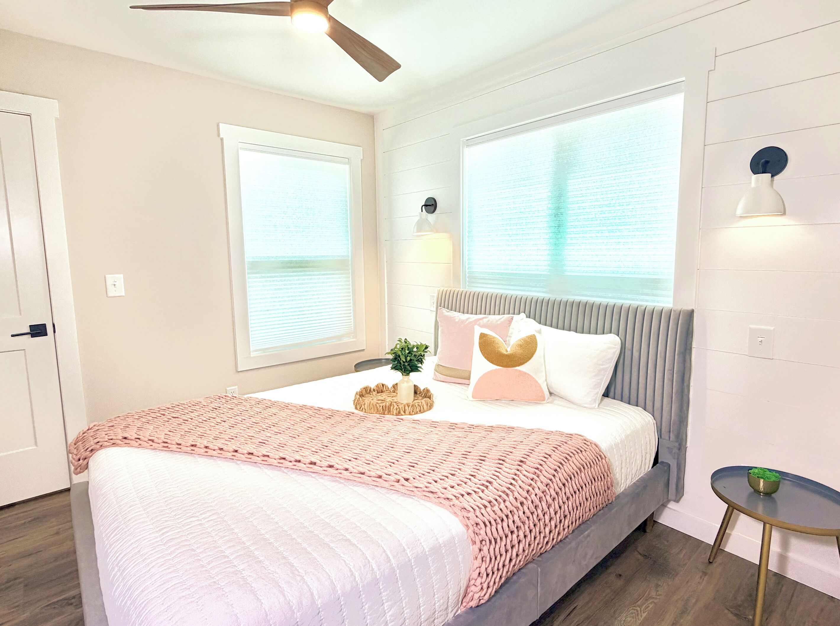 A bedroom with a bed, a ceiling fan, two windows, a bedside table, and a nightstand.