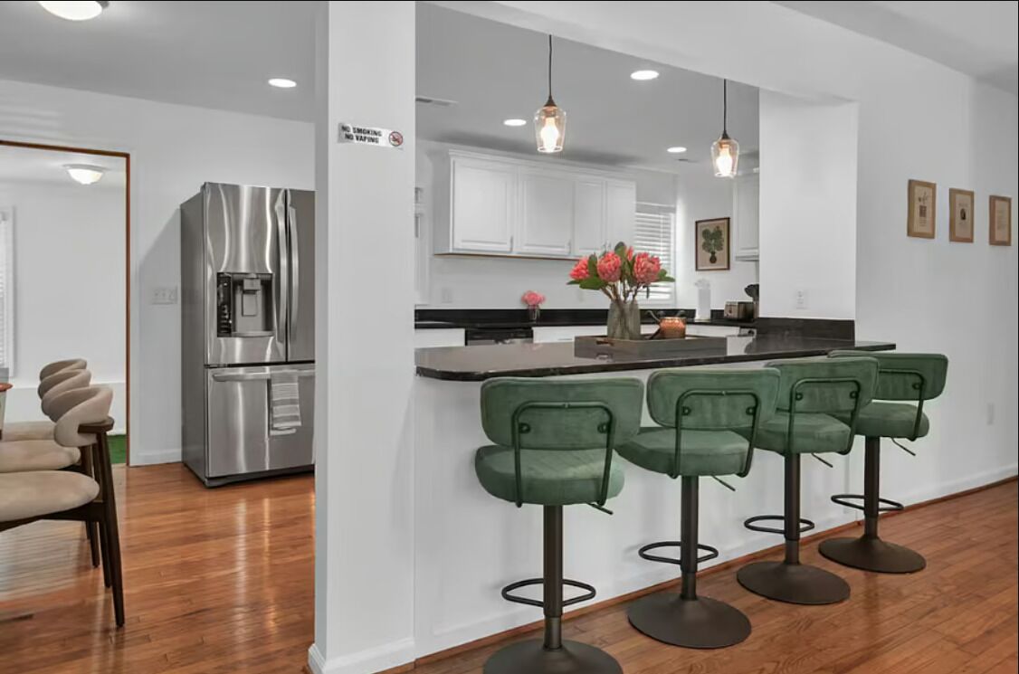 Une cuisine moderne avec un réfrigérateur en acier inoxydable, un coin bar doté de tabourets verts et un îlot central agrémenté d’un vase de fleurs.