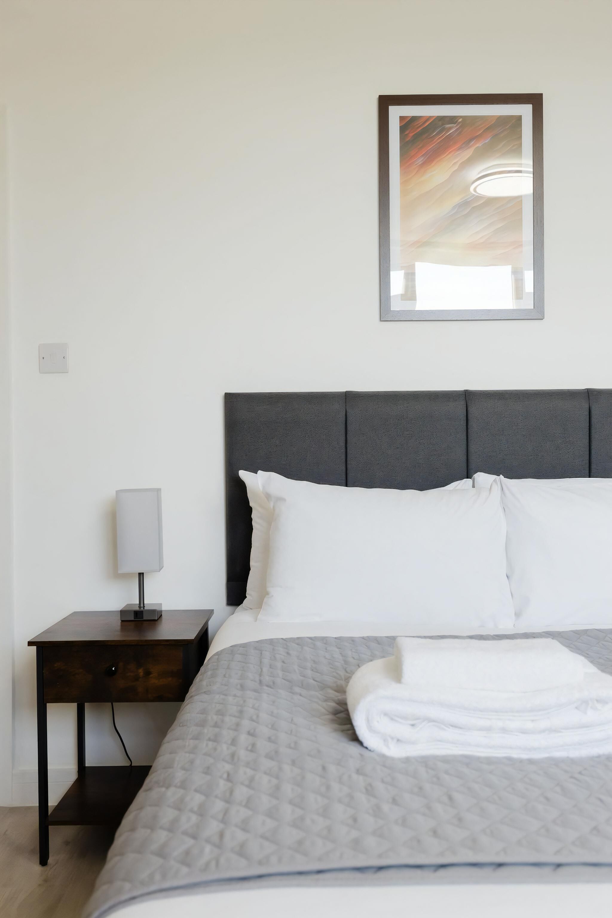 Una cama bien hecha con ropa de cama blanca, un cabecero gris y una lámpara de noche sobre una mesita de madera.