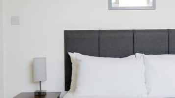 Una cama bien hecha con ropa de cama blanca, un cabecero gris y una lámpara de noche sobre una mesita de madera.