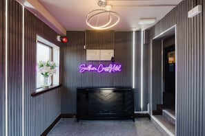 A hotel reception area with a neon sign, a reception desk, a window with flowers, and a modern light fixture.