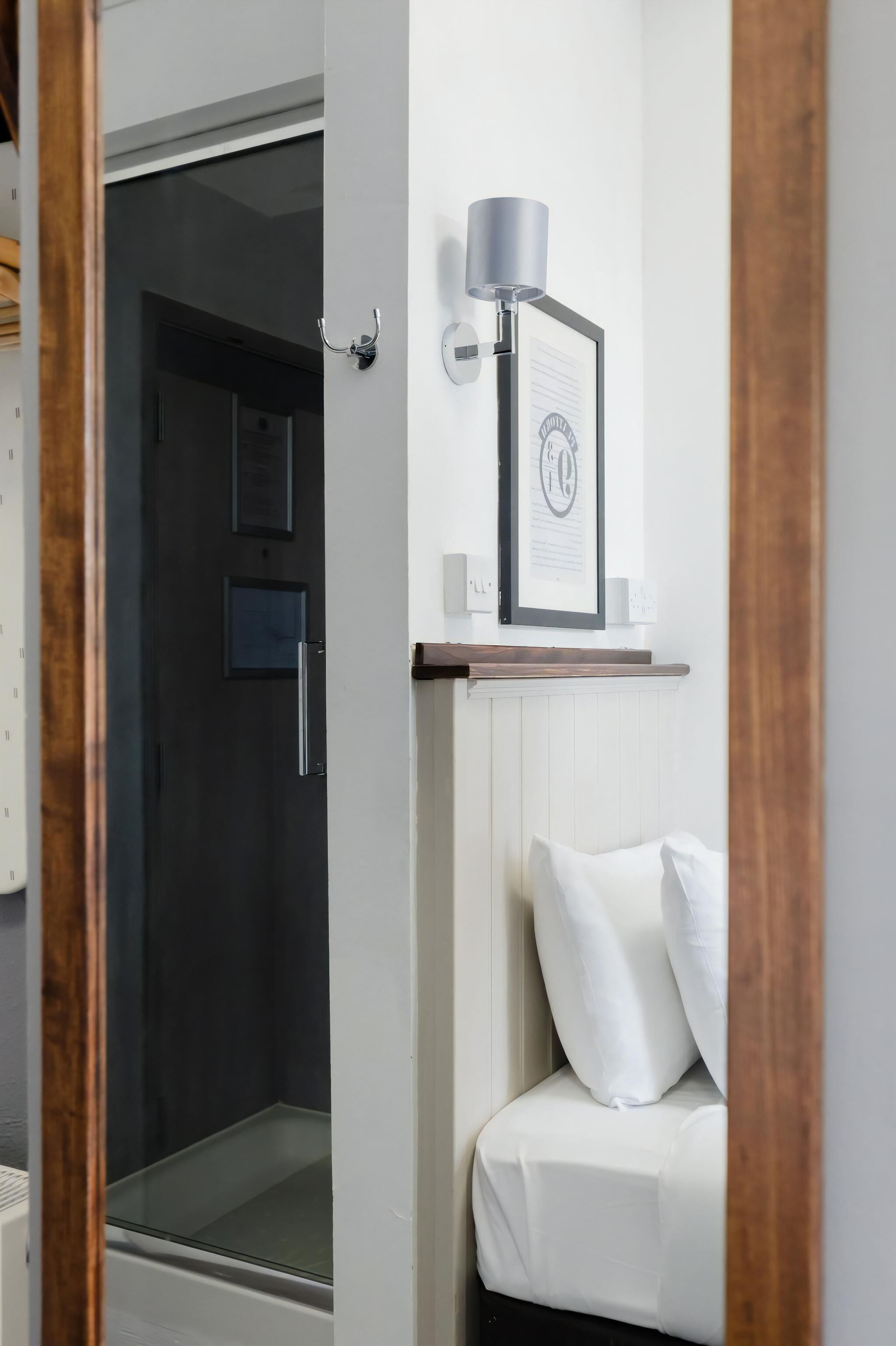 A hotel room with a bed, a framed certificate on the wall, a bedside lamp, and a door leading to a shower area.