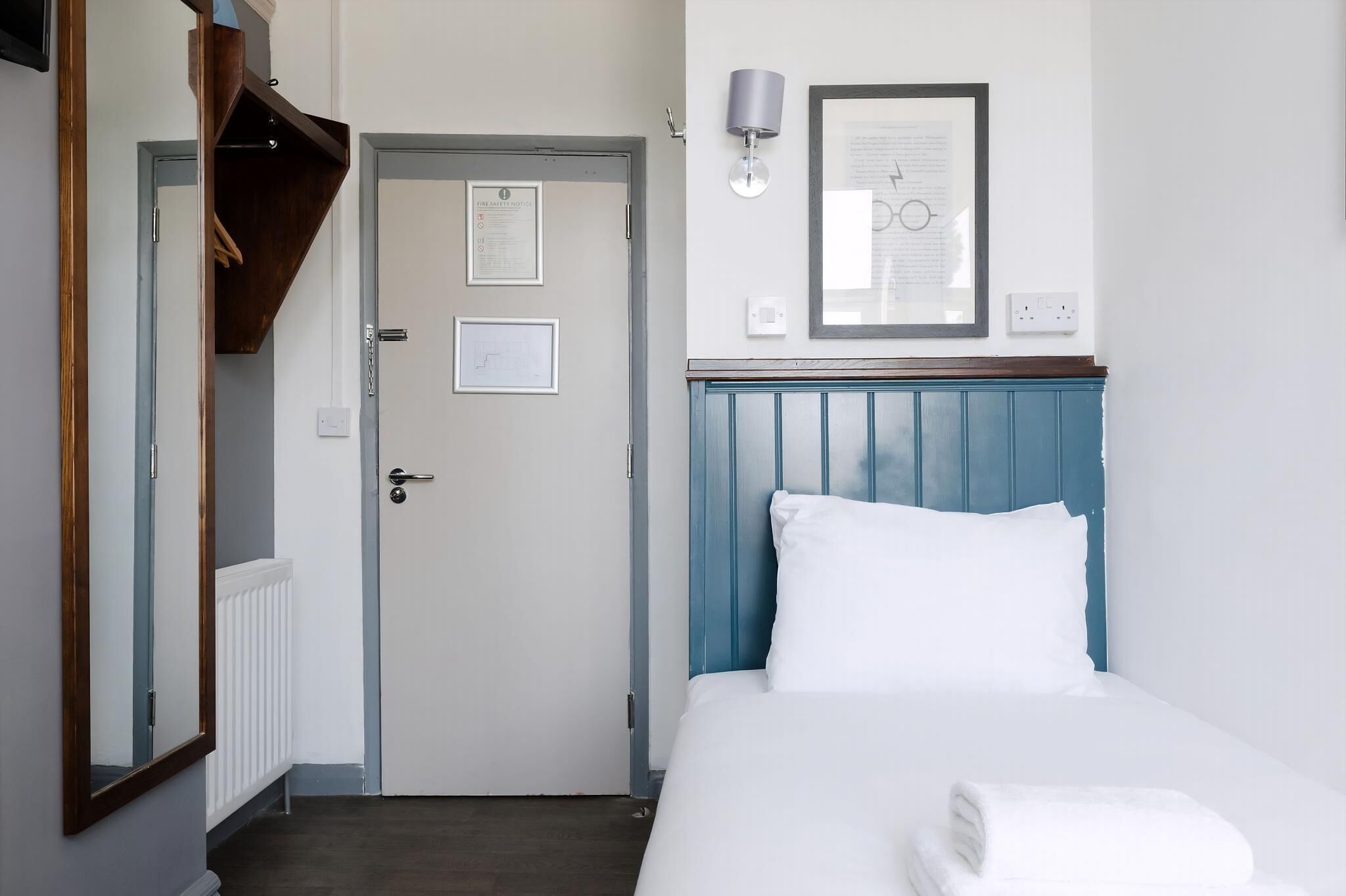 A hotel room with a bed, a door, a mirror, a wall-mounted lamp, and a framed document on the wall.