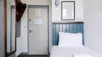 A hotel room with a bed, a door, a mirror, a wall-mounted lamp, and a framed document on the wall.