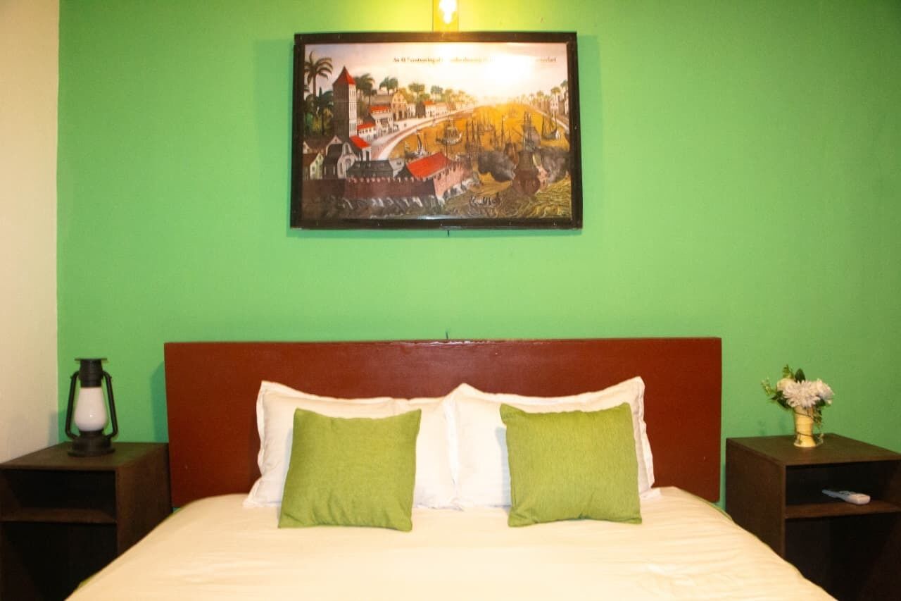 Een bed met witte en groene kussens, een houten hoofdbord, een nachtkastje met een lamp, en een ingelijste foto aan de muur.