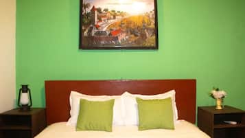 Een bed met witte en groene kussens, een houten hoofdbord, een nachtkastje met een lamp, en een ingelijste foto aan de muur.