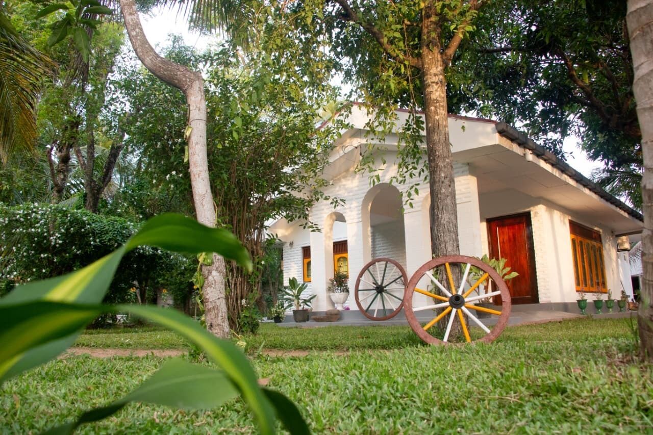 A building with a white facade, arched windows, and a red door. There is a large wheel in the foreground, and the surrounding area is lush with greenery.