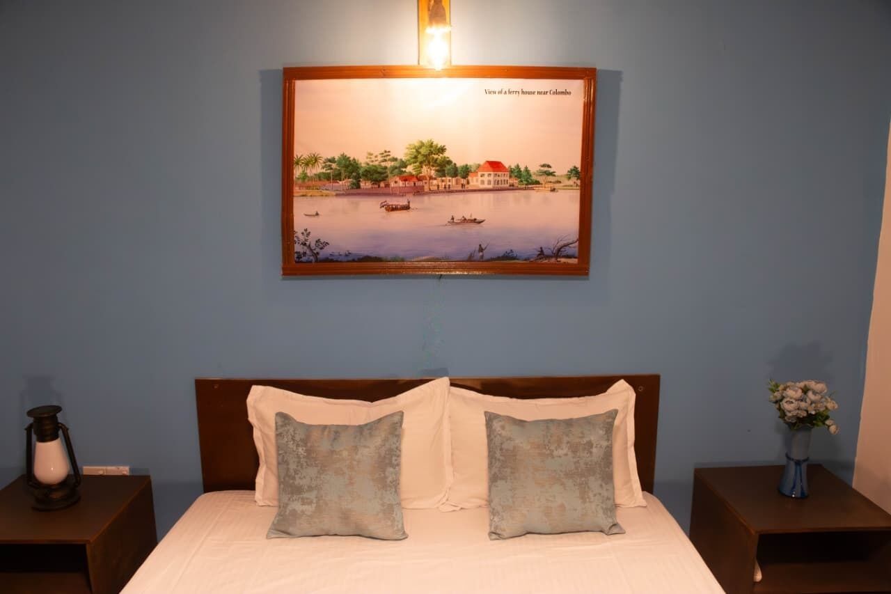 A bed with two pillows, a nightstand with a lamp, and a framed picture of a house near Colombo.