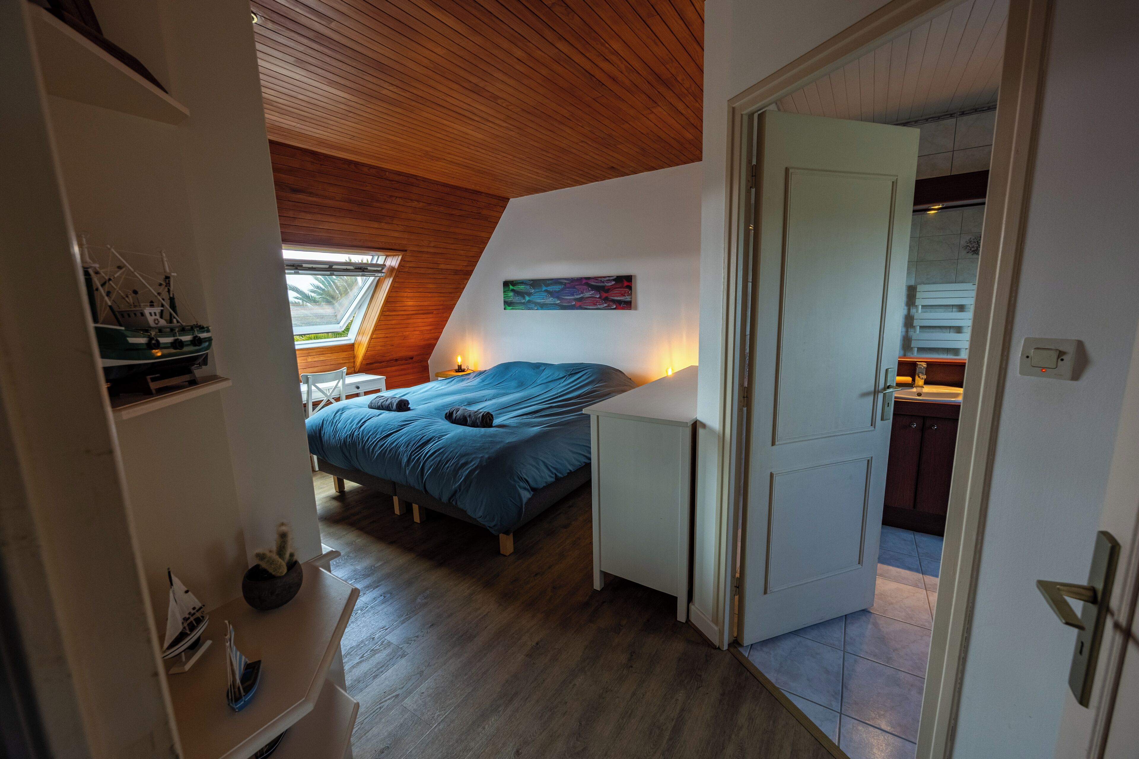 A bedroom with a sloped ceiling, a bed with blue bedding, a small table, and a wall-mounted shelf with decorative items.