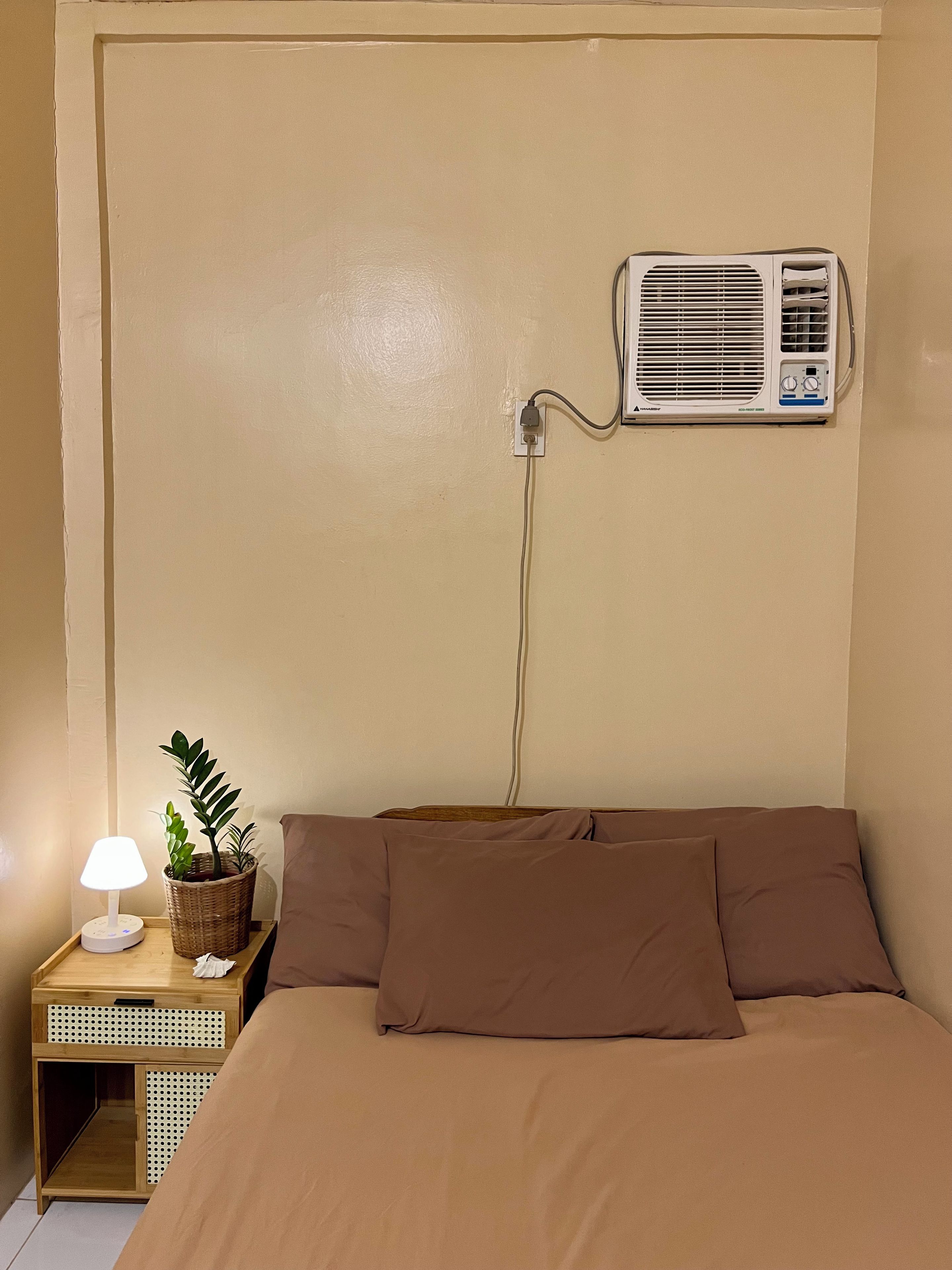 A bedroom with a bed, a bedside table with a lamp, a potted plant, and an air conditioning unit mounted on the wall.