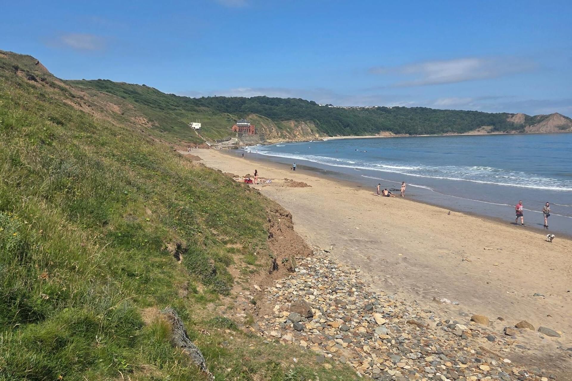 A coastal beach with a dirt path leading to the water, a few people, and a building in the background.