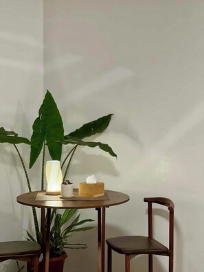 A round wooden table with a white candle and a tissue box, flanked by two chairs and a potted plant.