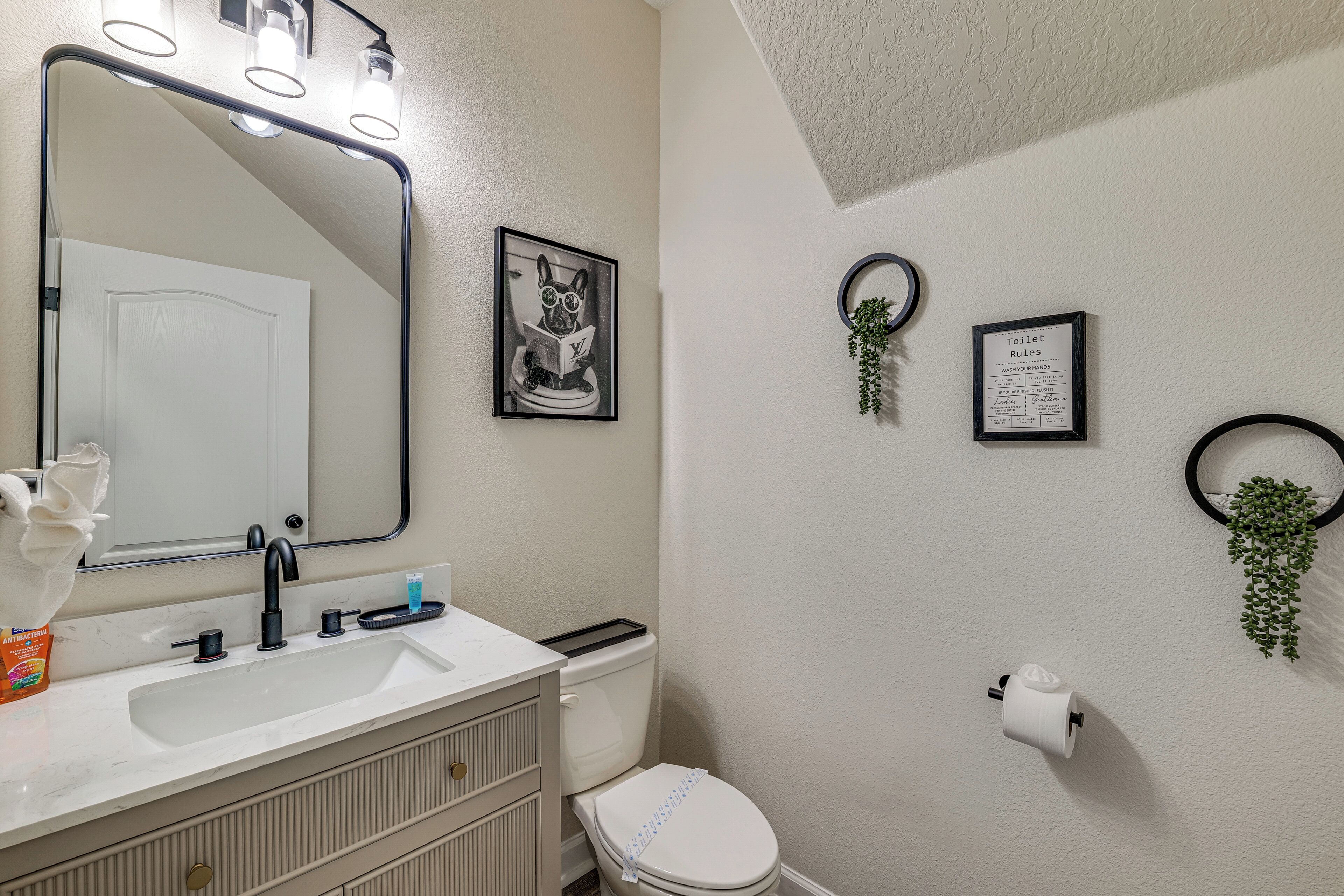 A bathroom with a white sink, mirror, and toilet. There are framed pictures on the wall, a toilet paper holder, and a small plant.