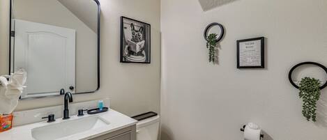 A bathroom with a white sink, mirror, and toilet. There are framed pictures on the wall, a toilet paper holder, and a small plant.