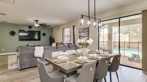 A dining area with a table set for four, a gray sofa, a flat-screen TV, and a ceiling fan. - New Remodeled Championsgate Pool Home 5br/5ba (Davenport)