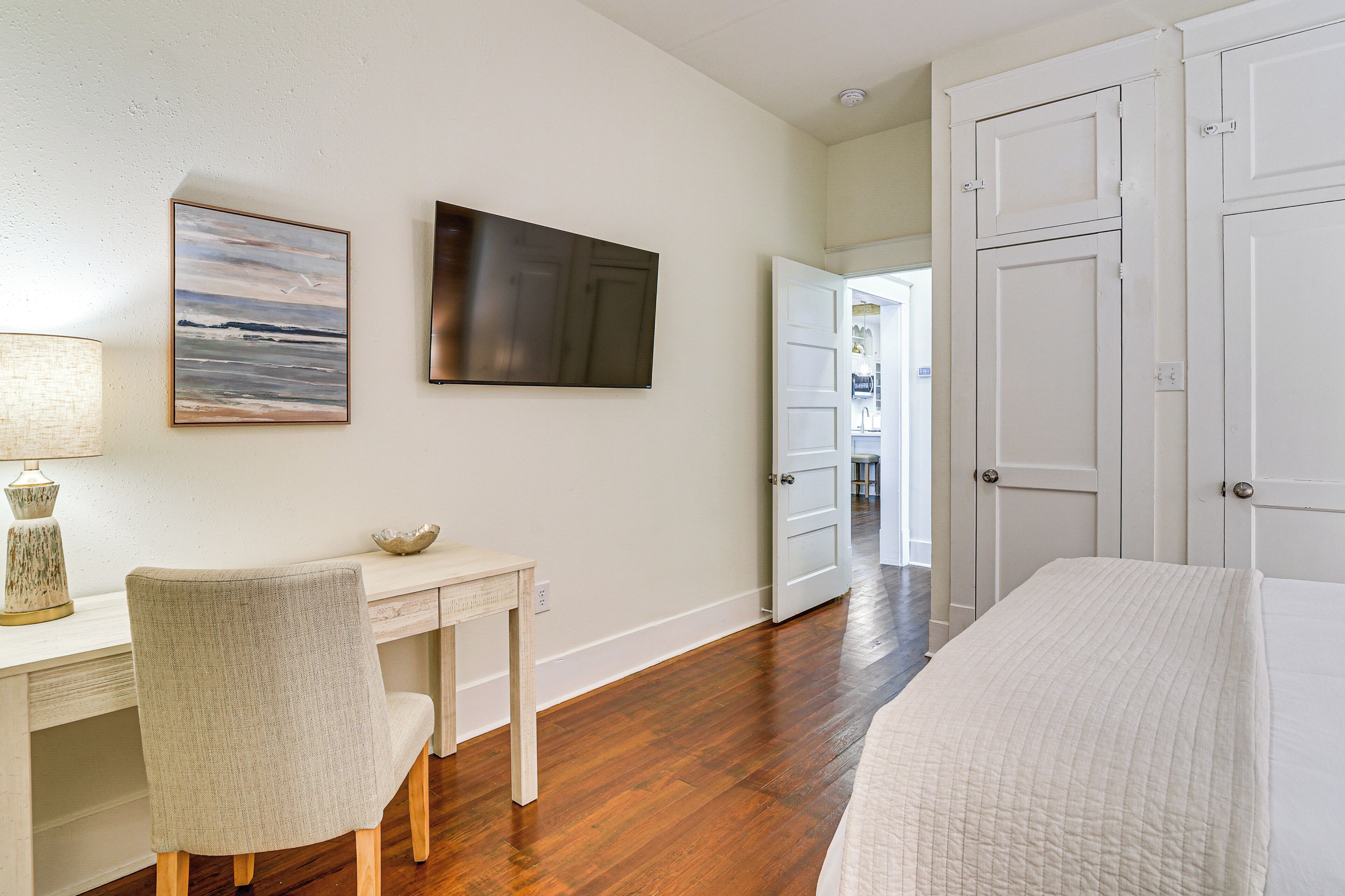 A bedroom with a bed, a desk with a chair, a painting, and a door leading to another room.