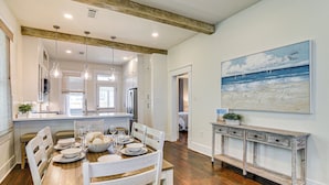 A dining area with a wooden table set for four, white chairs, a white console table, and a painting of a beach scene on the wall. - 1 Block to Beach: Fire Pit, Outdoor Kitchen, Bikes (Galveston)