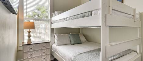 A bunk bed with a nightstand and lamp, a window with blinds, and a painting on the wall.