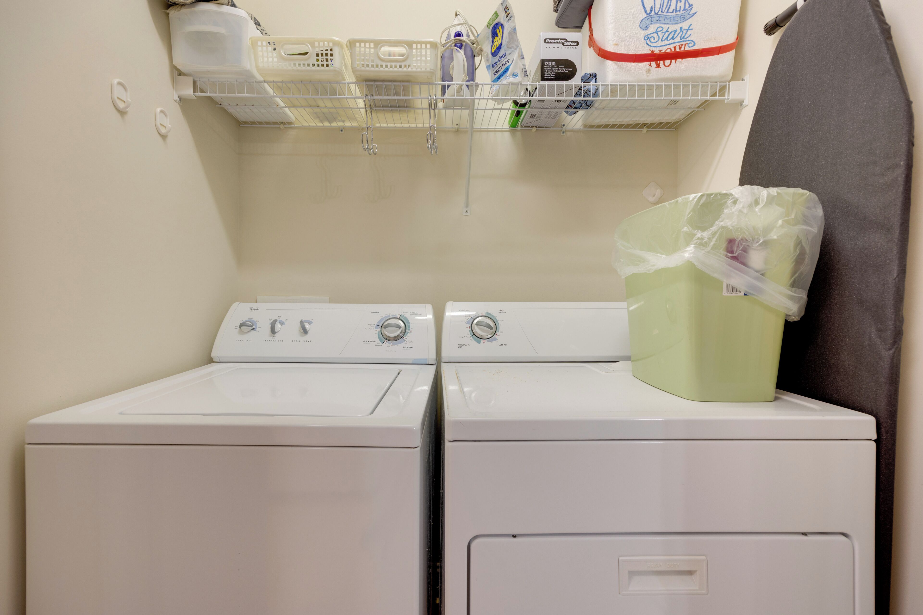 A laundry area with two white washers and dryers, a shelf with various laundry supplies, and a plastic bag on the dryer.