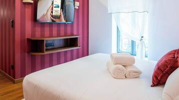 Quarto de hotel com uma cama, uma televisão fixada na parede, uma janela com cortinas e uma prateleira de madeira.