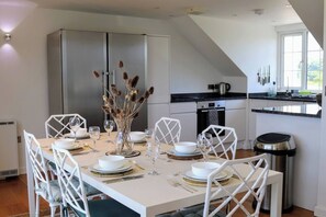 Un coin repas avec une table blanche dressée pour six personnes, des chaises blanches et un vase contenant des plantes séchées.