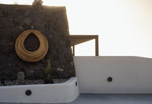 A stone wall with a woven basket, a cactus, and a white planter with black buttons. - Lithora Suites (Oia)