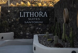 Une enseigne indiquant « LITHORA SUITES SANTORINI », avec un mur de pierres et des cactus au premier plan.