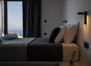 A hotel room with a bed, a nightstand, a wall-mounted lamp, and a view of the sea. - Lithora Suites (Oia)