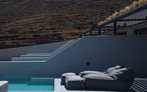 A modern outdoor pool area with a built-in seating area and a view of a hilly landscape. - Lithora Suites (Oia)