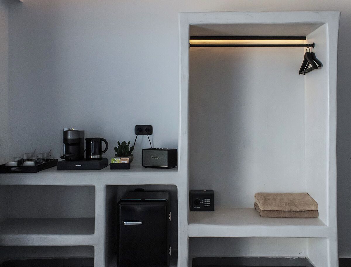 Habitación moderna con pared blanca, repisa con cafetera, planta, radio, pequeño calentador, maleta y toalla doblada.
