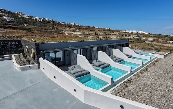 A modern outdoor pool area with multiple water slides and lounge chairs, surrounded by a pebble beach and a hillside view. - Lithora Suites (Oia)