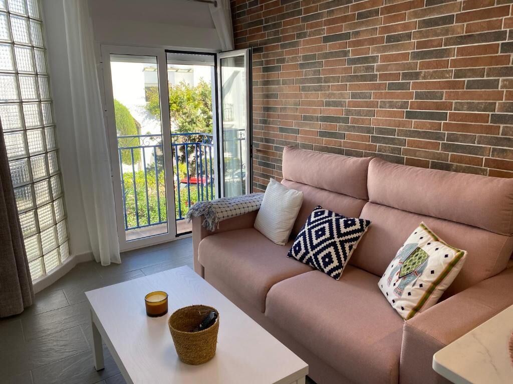 A modern living room with a brick wall, a sofa with patterned cushions, a white table, and a view through a glass door to an outdoor balcony.