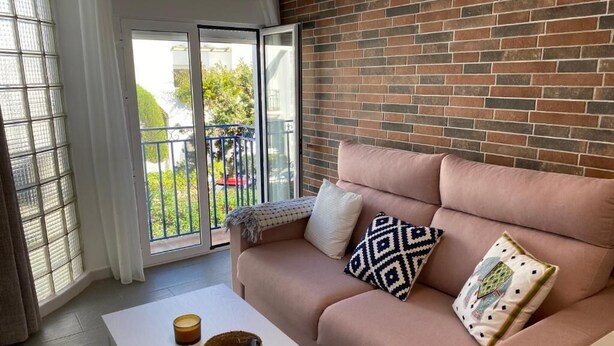 Ein modernes Wohnzimmer mit Backsteinwand, einer Couch mit gemusterten Kissen, einem weißen Tisch und Blick durch eine Glastür auf einen Außenbalkon.